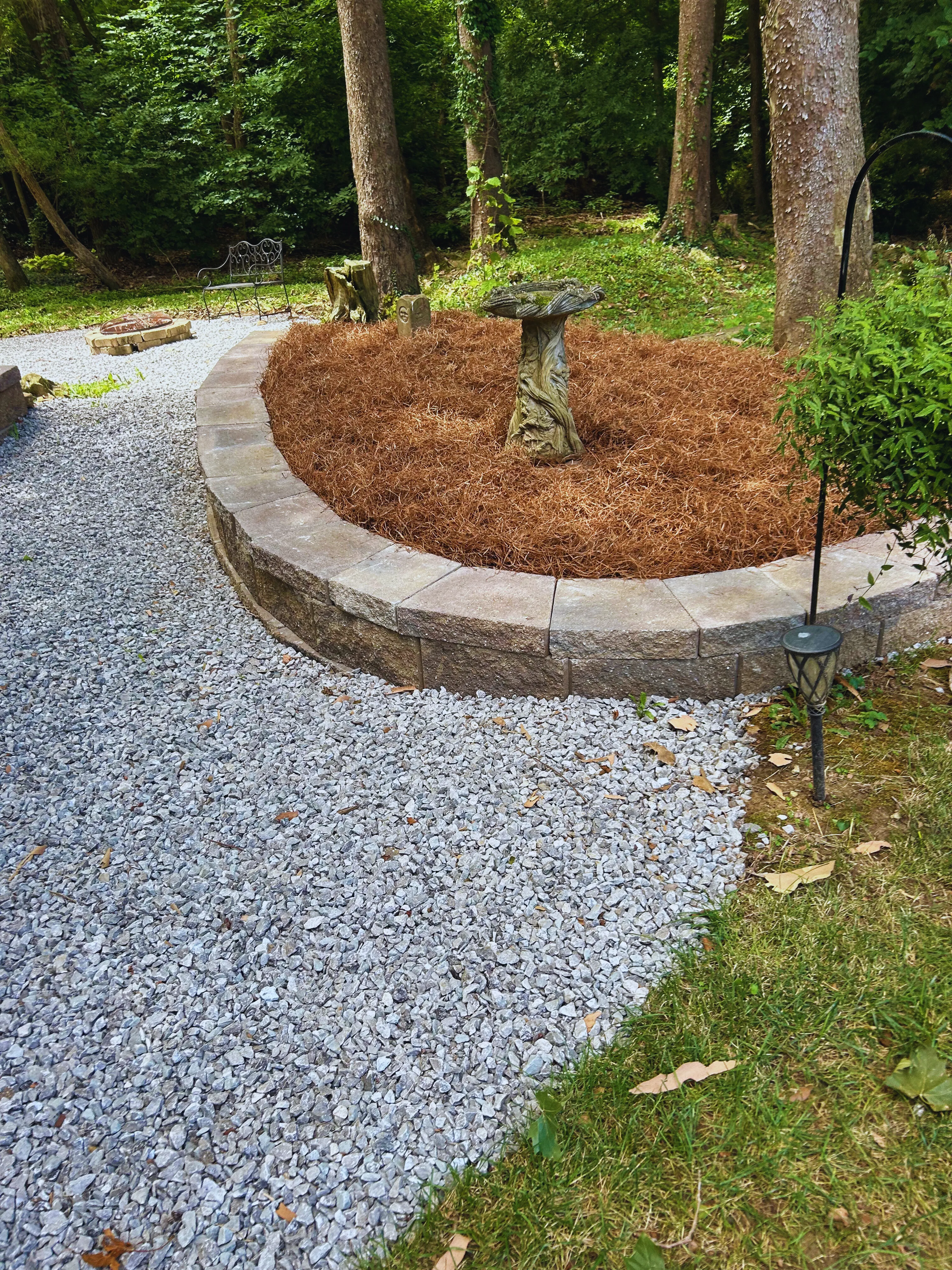 Curved stone retaining wall around a tree, with a garden statue and pine straw mulch